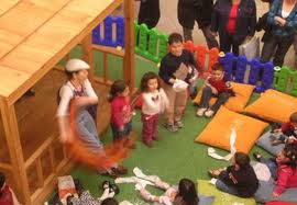 Ahşap oyun evi çevresinde minderler üzerindeki çocuklar ve etkinlik yürütücüsü