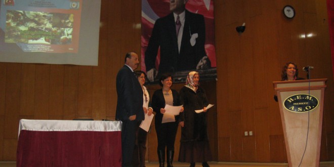 Salon sahnesinde belge tutan katılımcılar ve arkadaki büyük portre