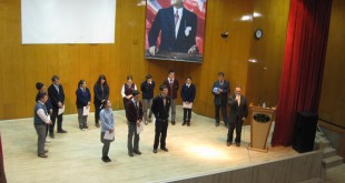 Okul salonunda sahnede sıralanmış öğrenciler ve arkadaki büyük portre