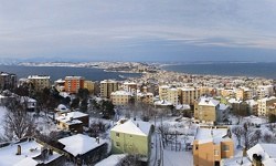 Karla kaplı çatılar ve arkada deniz görünen şehir manzarası