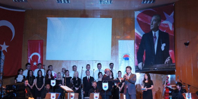 Sahnede saz heyeti ve koro, arkada Türk bayrağı ve büyük portre