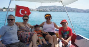 Bayraklı teknede oturan yetişkinler ve çocuklar, arkada kıyı şehri