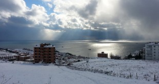Karla kaplı kıyı kasabasında bulutlar arasından süzülen güneş ışıklarıyla deniz manzarası
