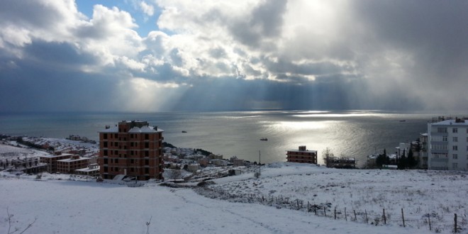 Karla kaplı kıyı kasabasında bulutlar arasından süzülen güneş ışıklarıyla deniz manzarası