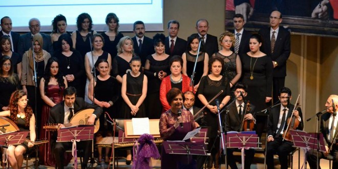 Sahnede koro ve çalgıcılar, önde mor kurdeleli mikrofon başında kadın