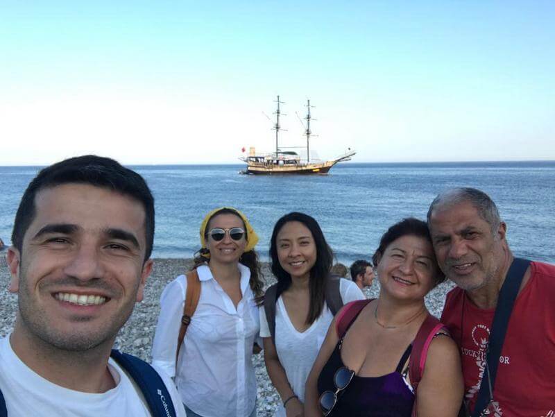 Deniz kenarında poz veren bir grup insanın arkasında yelkenli bir tekne görünüyor.