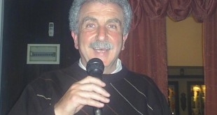 Mikrofon tutarak gülümseyen erkek, arkasında bordo perde