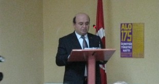 Bayrak yanındaki kürsüde konuşma yapan takım elbiseli erkek